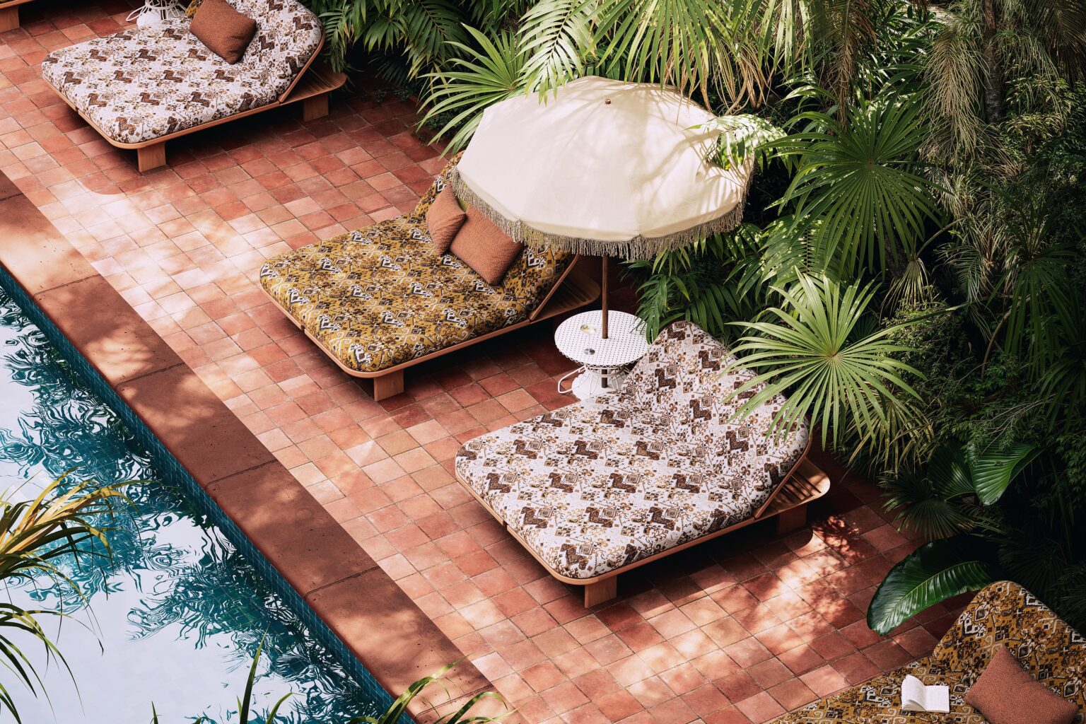Vue aérienne de trois daybeds en bois clair au bord d'une piscine sur un sol en tomettes terre cuite, recouverts de matelas à motifs ethniques dans des tons ocre, brun et blanc cassé avec coussins bouclés terracotta, séparés par une petite table ronde blanche sous un parasol écru à franges, entourés de palmiers et de végétation tropicale dense.